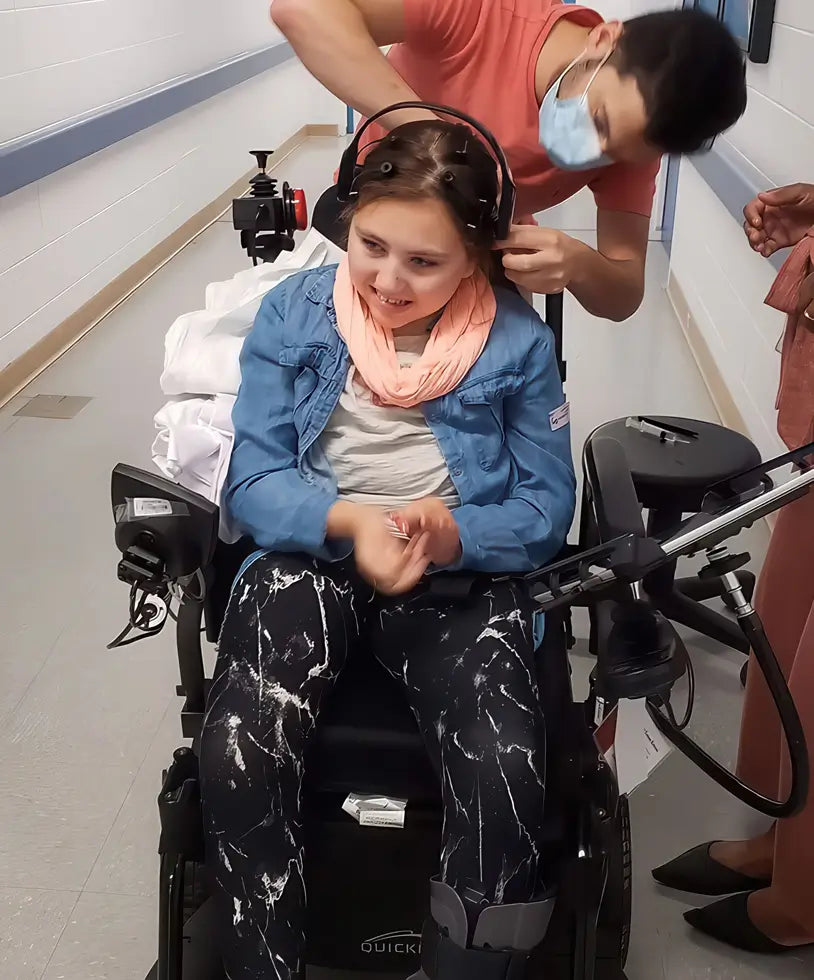 A girl sitting on a wheelchair while a researcher sets up an Emotiv Epoc X on her head